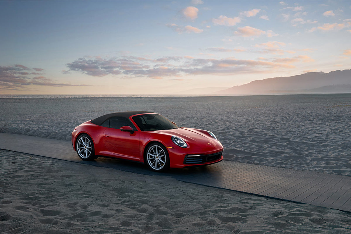 Red Porsche 911 Carrera cabriolet parked on a beach boardwalk at dusk with the ocean and mountains behind, by Niaz Uddin